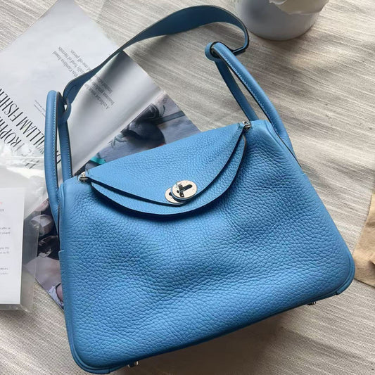 A light blue pebbled leather handbag with a silver-tone turn-lock closure, laid flat on a textured beige fabric surface with a magazine and a small flower arrangement in the background.
