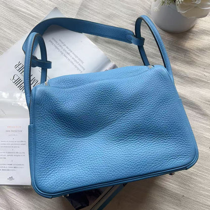 A light blue pebbled leather shoulder bag with a structured rectangular shape, resting on white papers with a small white flower in a vase in the top-left corner.