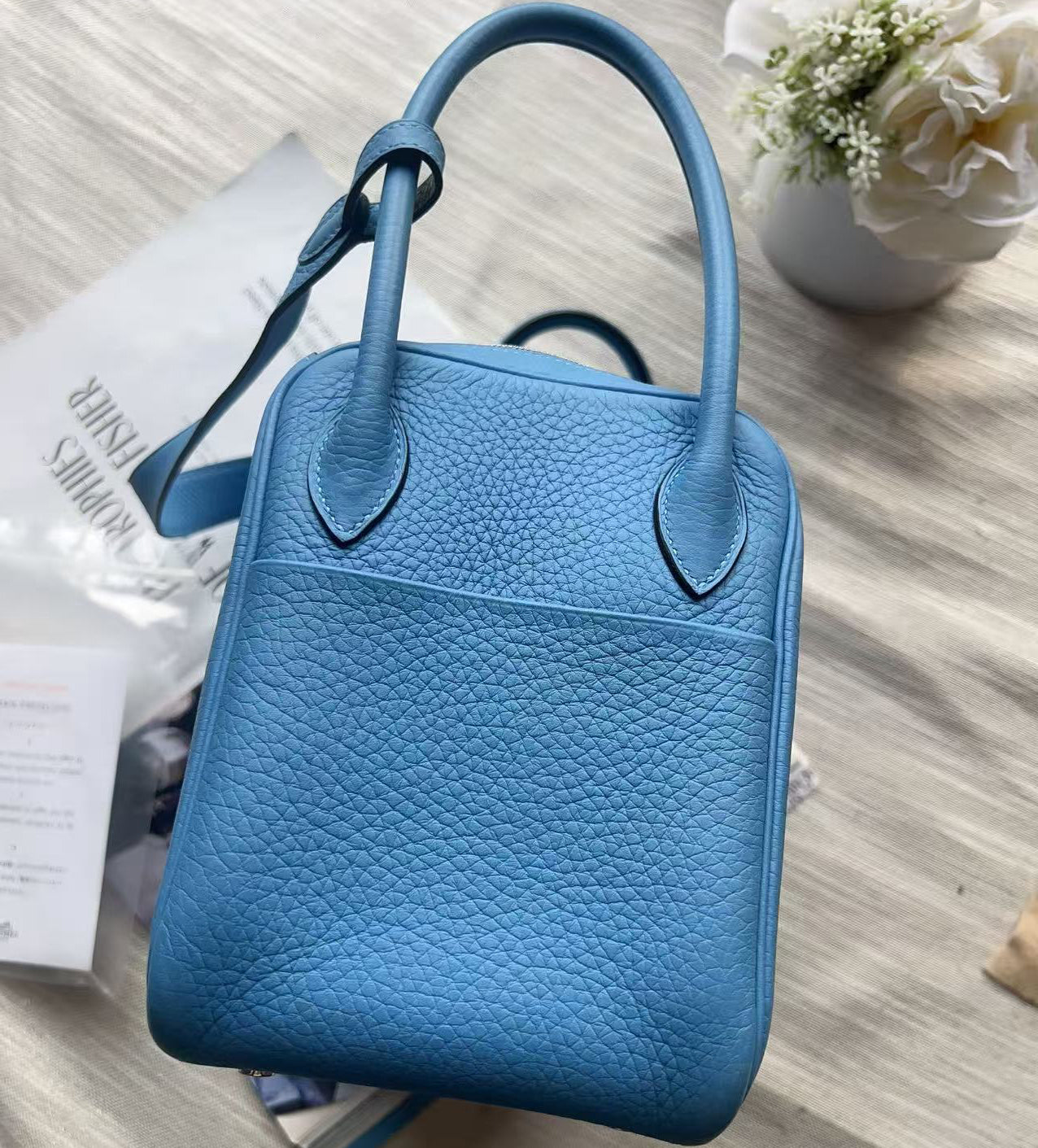 A light blue pebbled leather handbag with top handles, photographed from above on a light beige fabric surface with a white flower and shopping packaging nearby.