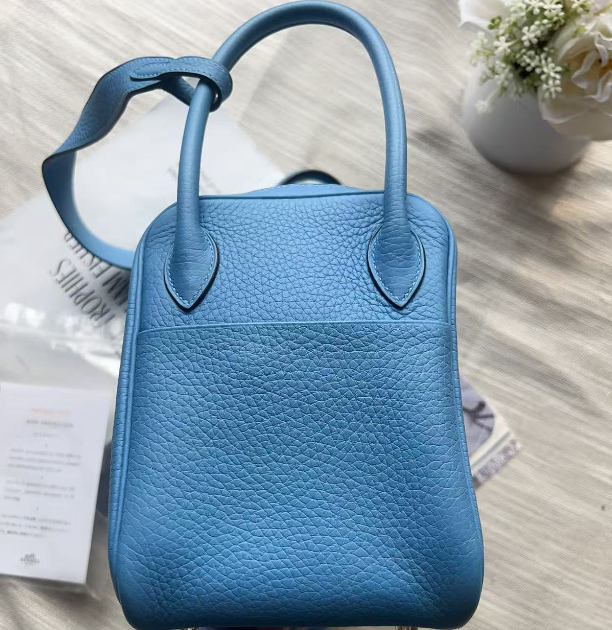 A blue pebbled leather handbag with double top handles, photographed from above on a light beige fabric surface with a small white flower arrangement in the background.