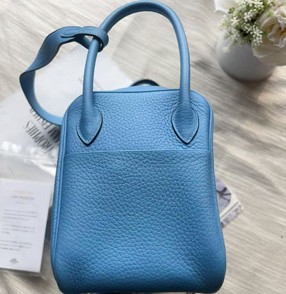 A blue pebbled leather handbag with double top handles, photographed from above on a light beige fabric surface with a small white flower arrangement in the background.