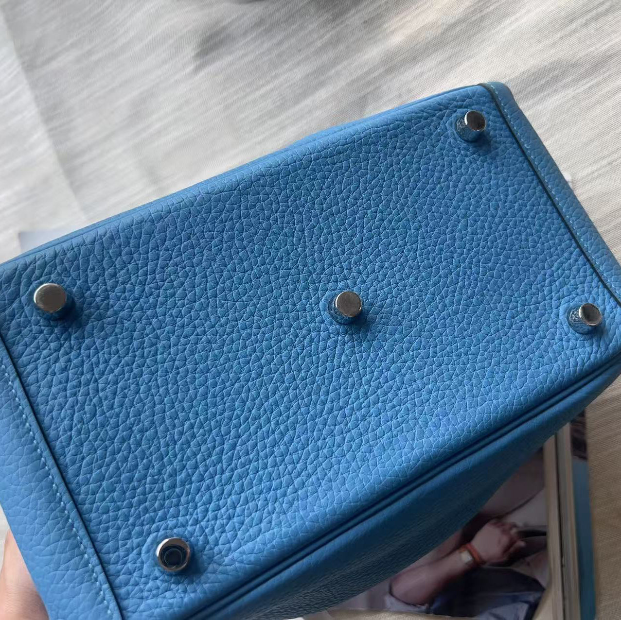 A close-up of a blue pebbled leather handbag bottom, showing five silver-toned metal studs on a light-colored wooden surface.