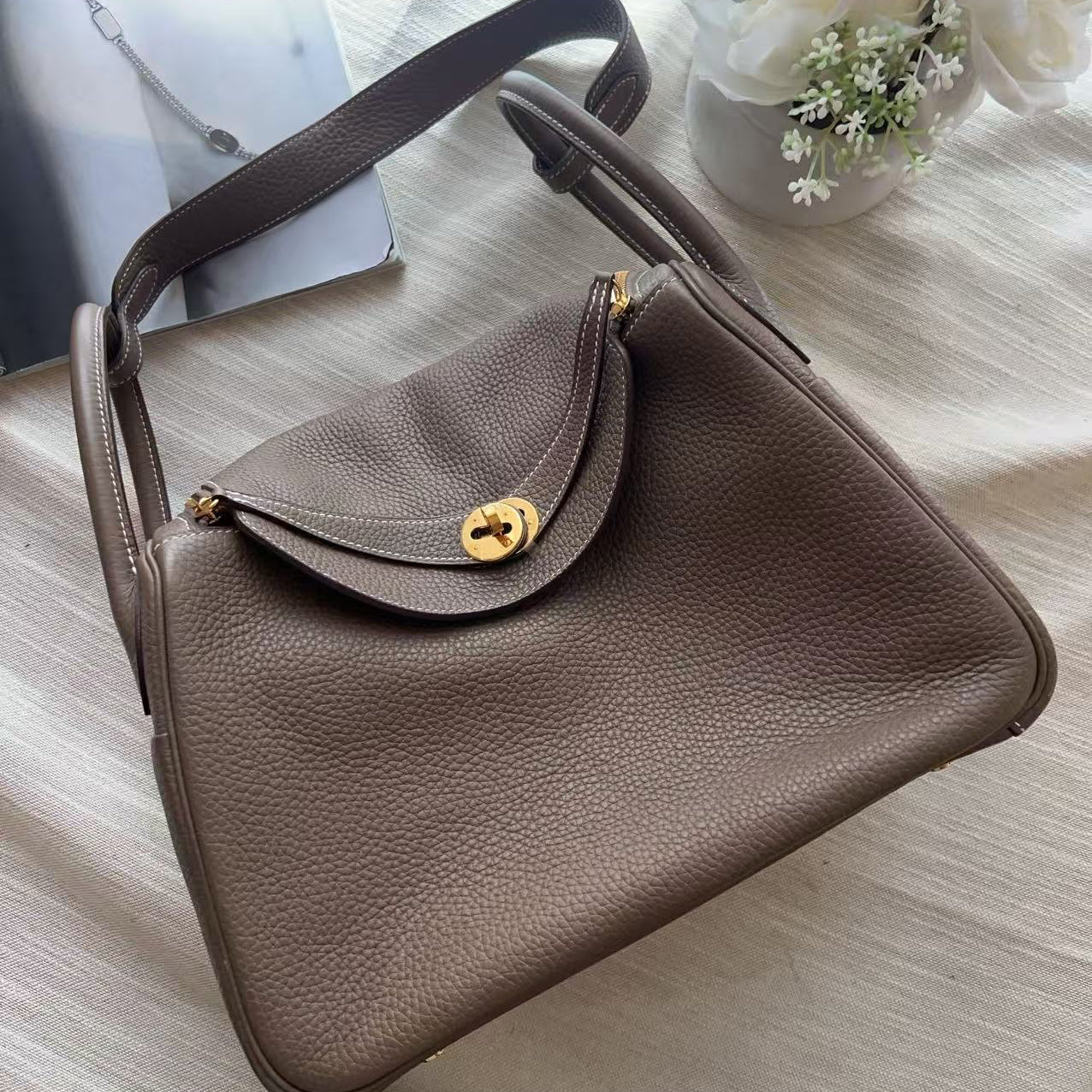A taupe brown pebbled leather handbag with a gold-tone turn-lock closure and strap, styled on a textured cream fabric with white flowers in the corner.