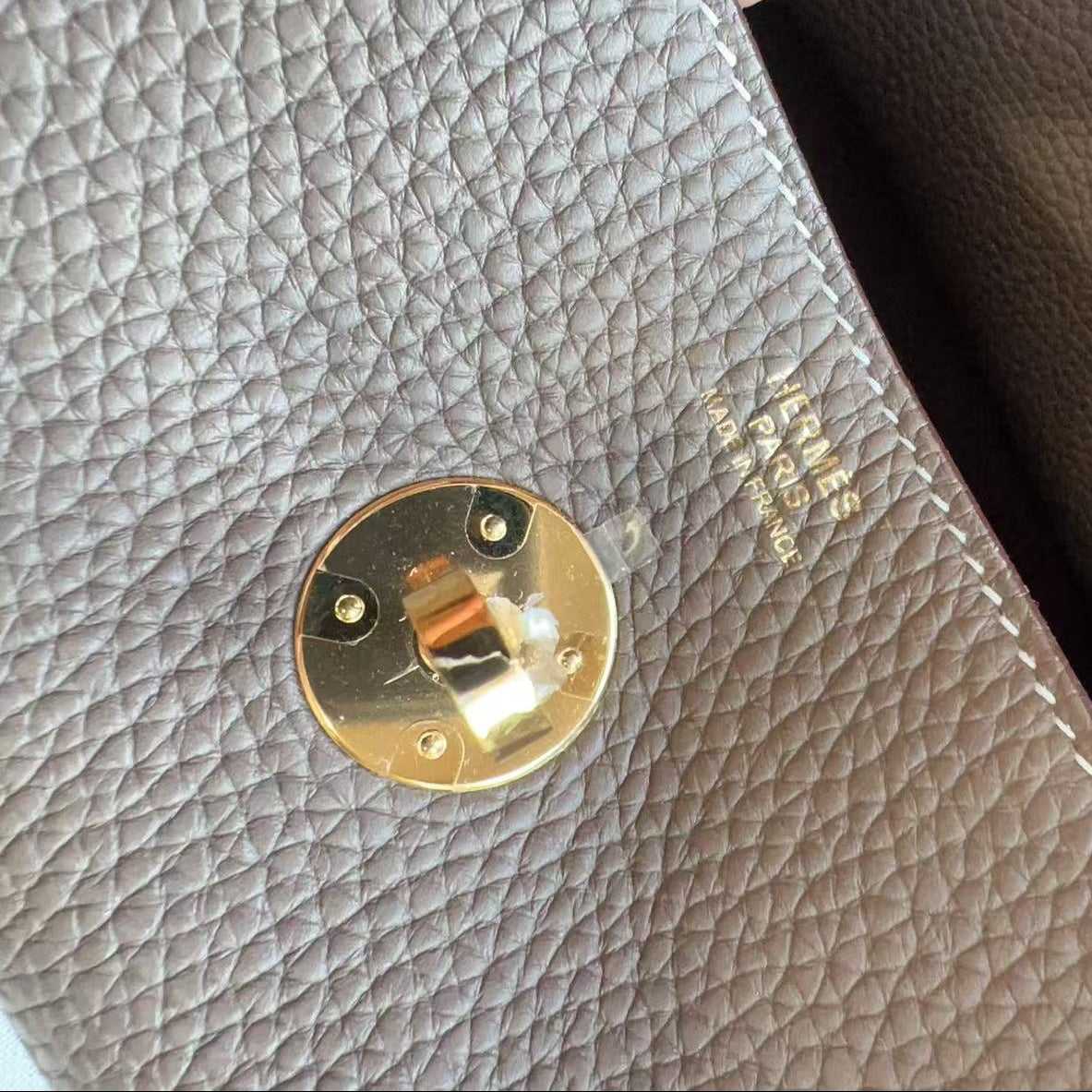 Close-up of a textured leather clutch or wallet showing a light taupe pebbled exterior with a gold-tone turn-lock closure and a dark brown interior lining.