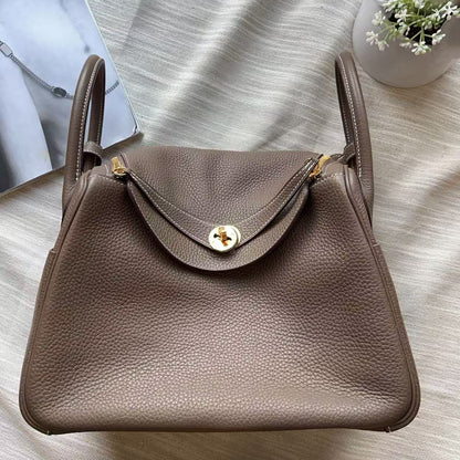 A taupe brown pebbled leather handbag with gold hardware, laid flat on a textured white fabric background with a magazine and small flowers as props.
