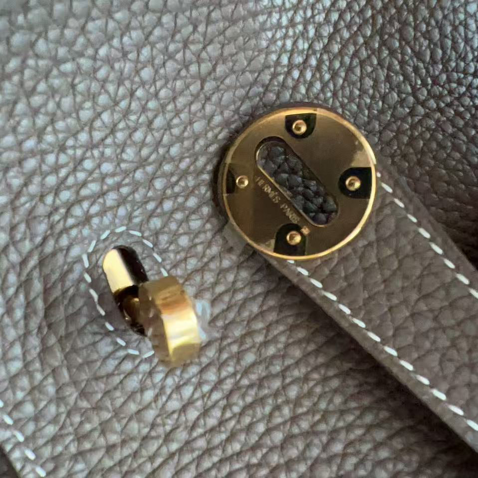 Close-up of a taupe pebbled leather bag with gold-tone hardware, showing a turn-lock clasp and a round lock plate, with a finger holding the strap on the right.