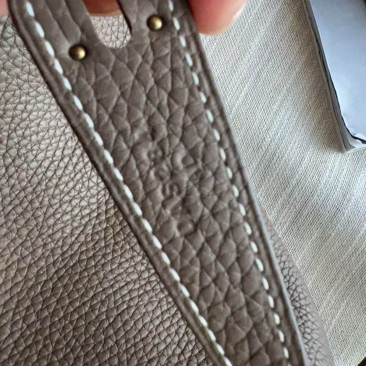 Close-up detail of a taupe textured leather handbag strap with white contrast stitching, gold-tone rivets, and a gold clasp, held by a hand over a light-colored fabric background.