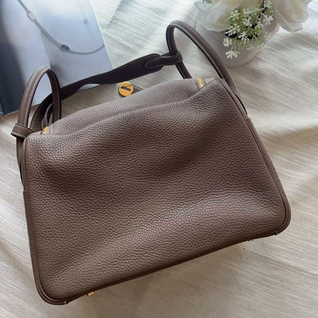 A taupe brown pebbled leather handbag with gold-tone hardware, laid flat on a textured white fabric surface next to white flowers.