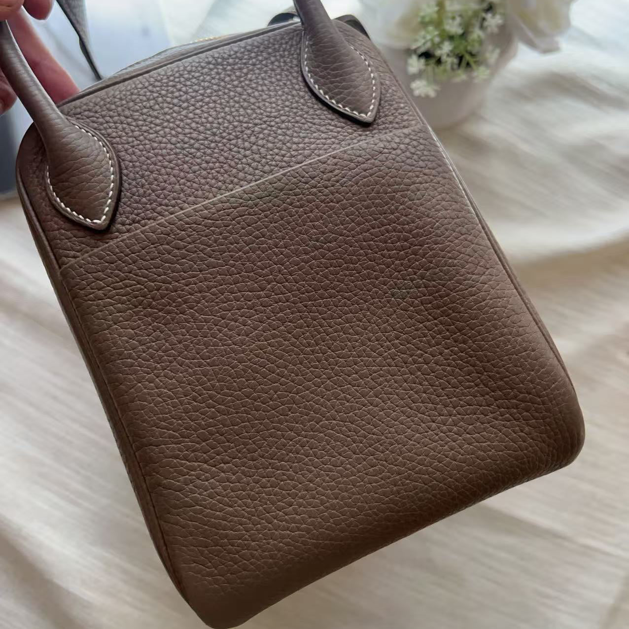 A taupe brown pebbled leather handbag with double top handles, held by a hand over a soft white fabric background with white flowers.