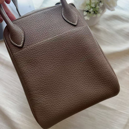 A taupe brown pebbled leather handbag with double top handles, held by a hand over a soft white fabric background with white flowers.