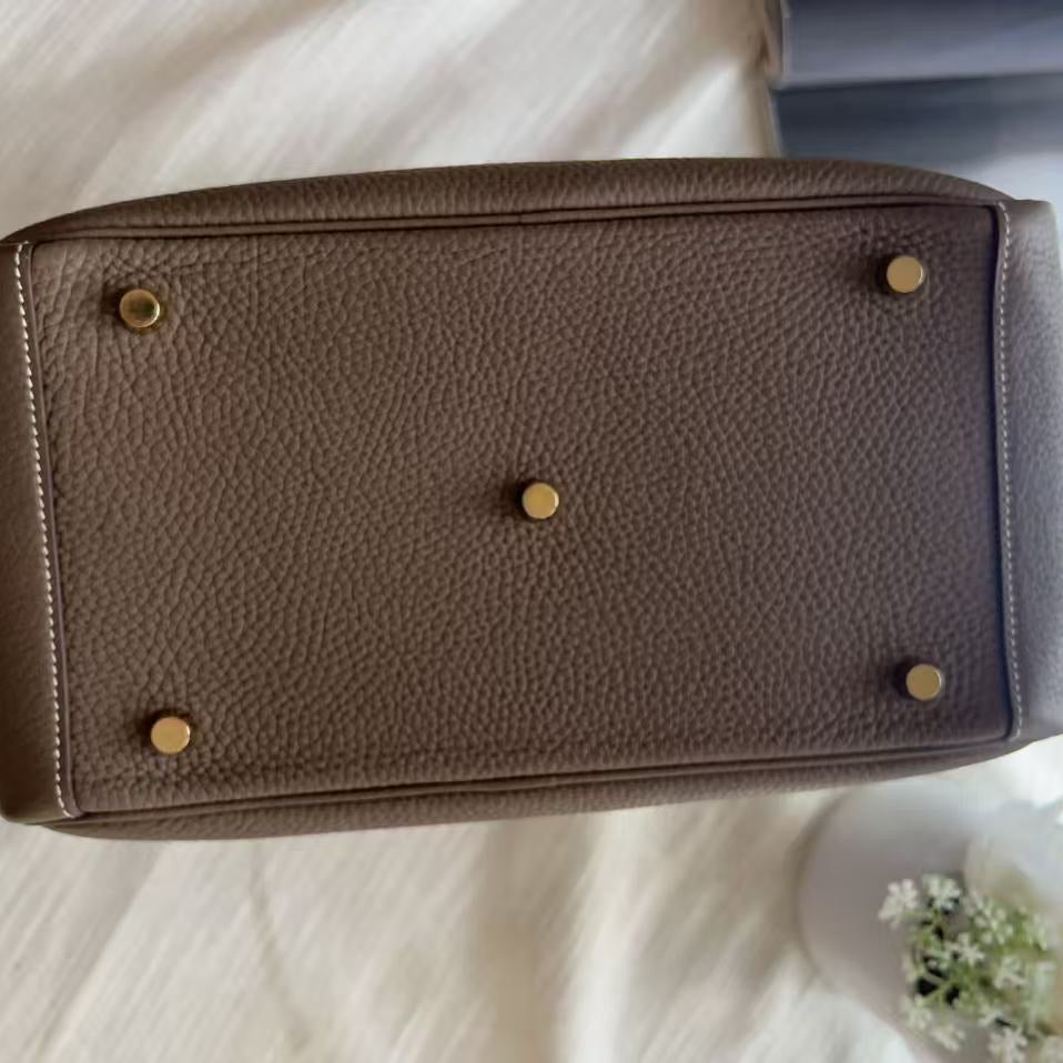 A top-down view of a brown pebbled leather handbag showing the bottom base with brass-tone corner and center studs, resting on a white fabric surface.