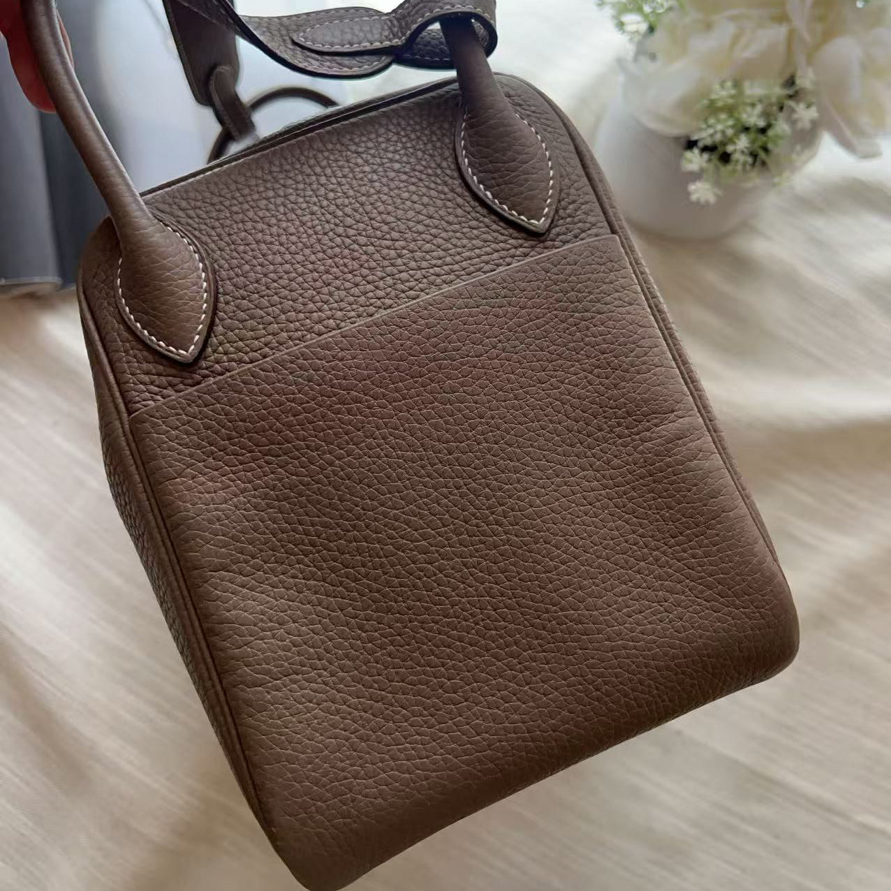A taupe brown pebbled leather handbag with top handles, held by a hand over a cream-colored fabric background with white flowers.