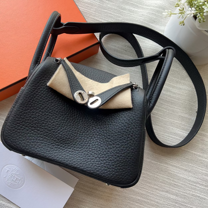 A black pebbled leather shoulder bag with a beige leather accent and silver-tone hardware, displayed on a light wood surface next to an orange Hermès box and a white card.