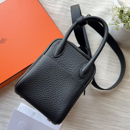 A black pebbled leather top-handle handbag with a matching shoulder strap, styled on a light wood surface next to an orange box and white flowers.