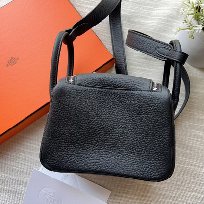 A black pebbled leather handbag with silver-tone hardware, displayed on a light wood surface next to an orange Hermès box and a white card.