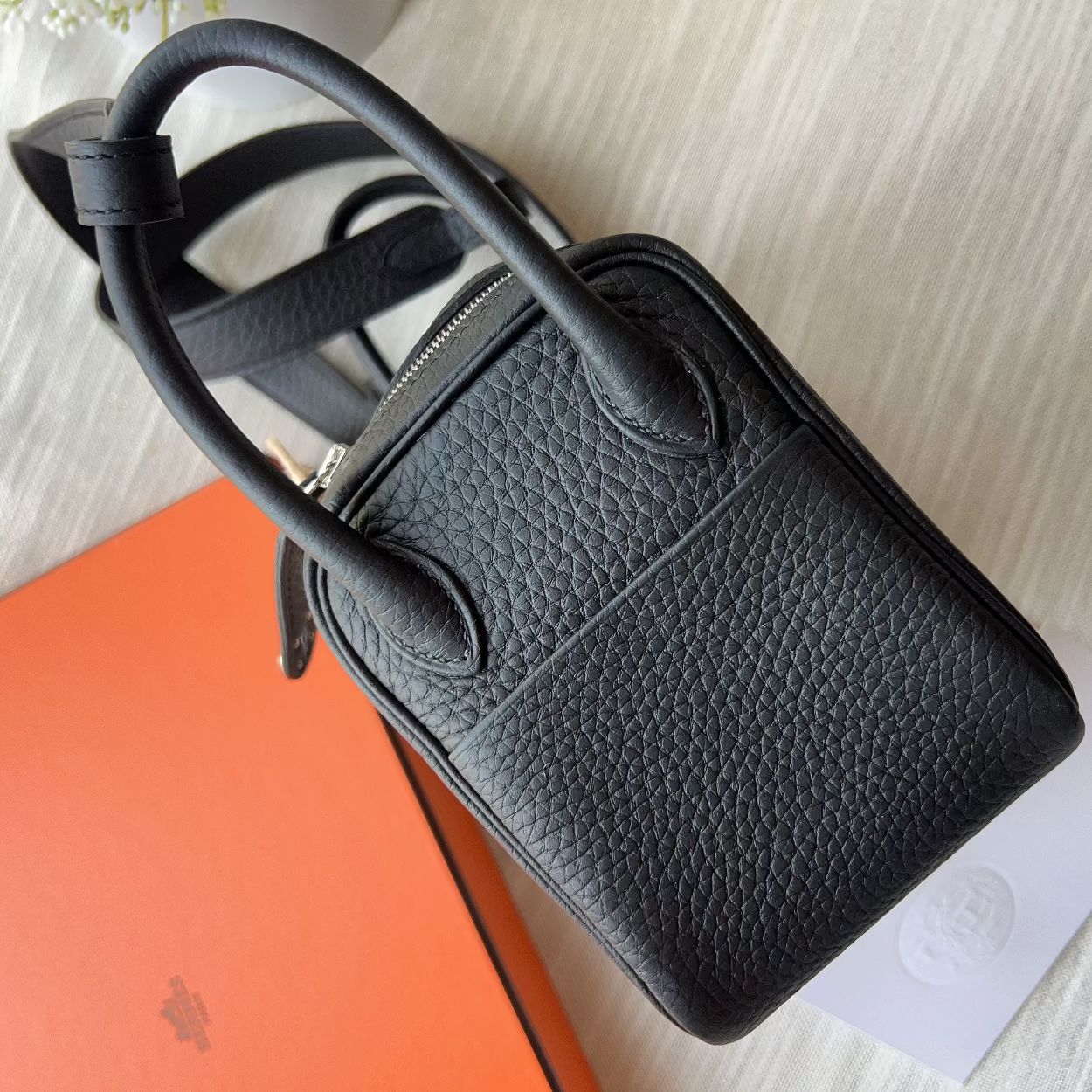 A black pebbled leather handbag with dual top handles and a zip-top closure, photographed on a light fabric surface with an orange box and white card underneath.