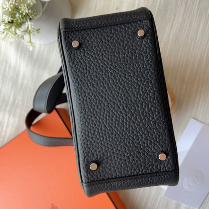 A black pebbled leather handbag shown from a top-down angle, resting on an orange box with a white card on a light beige fabric surface.