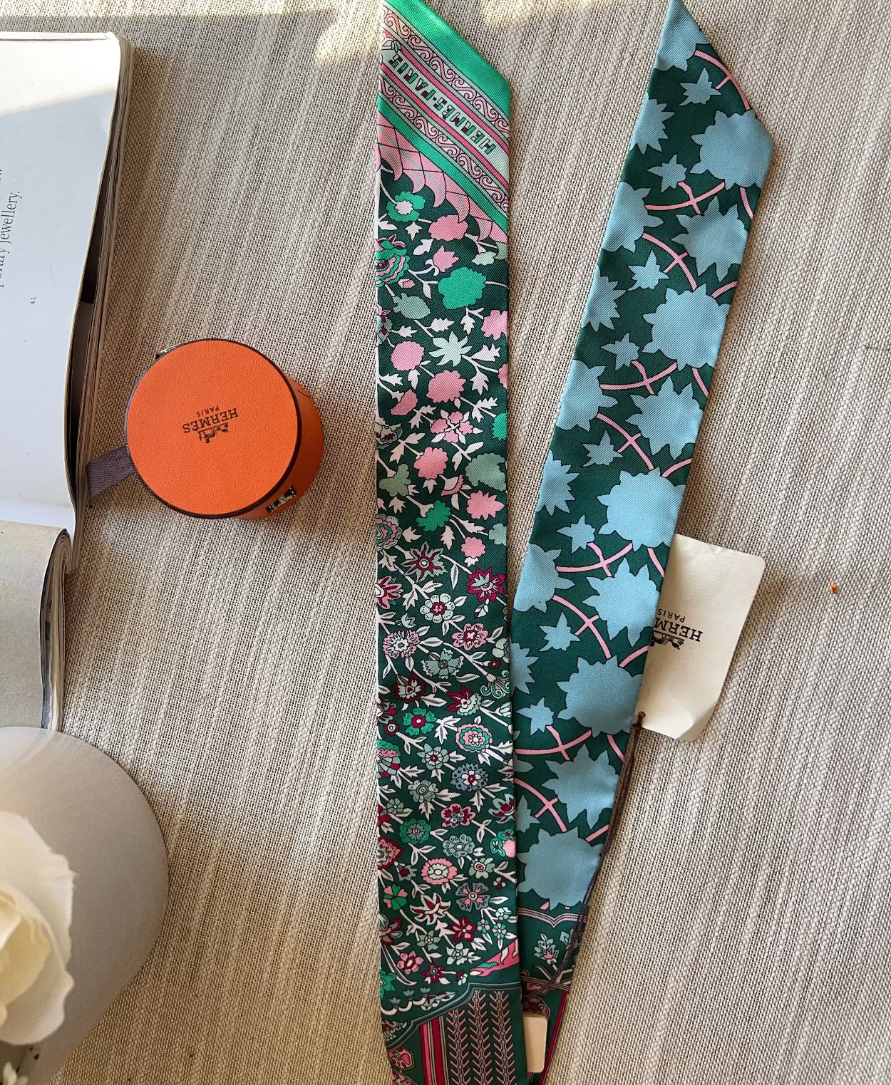 Two patterned scarves on a wooden surface with a small orange container and white flowers.