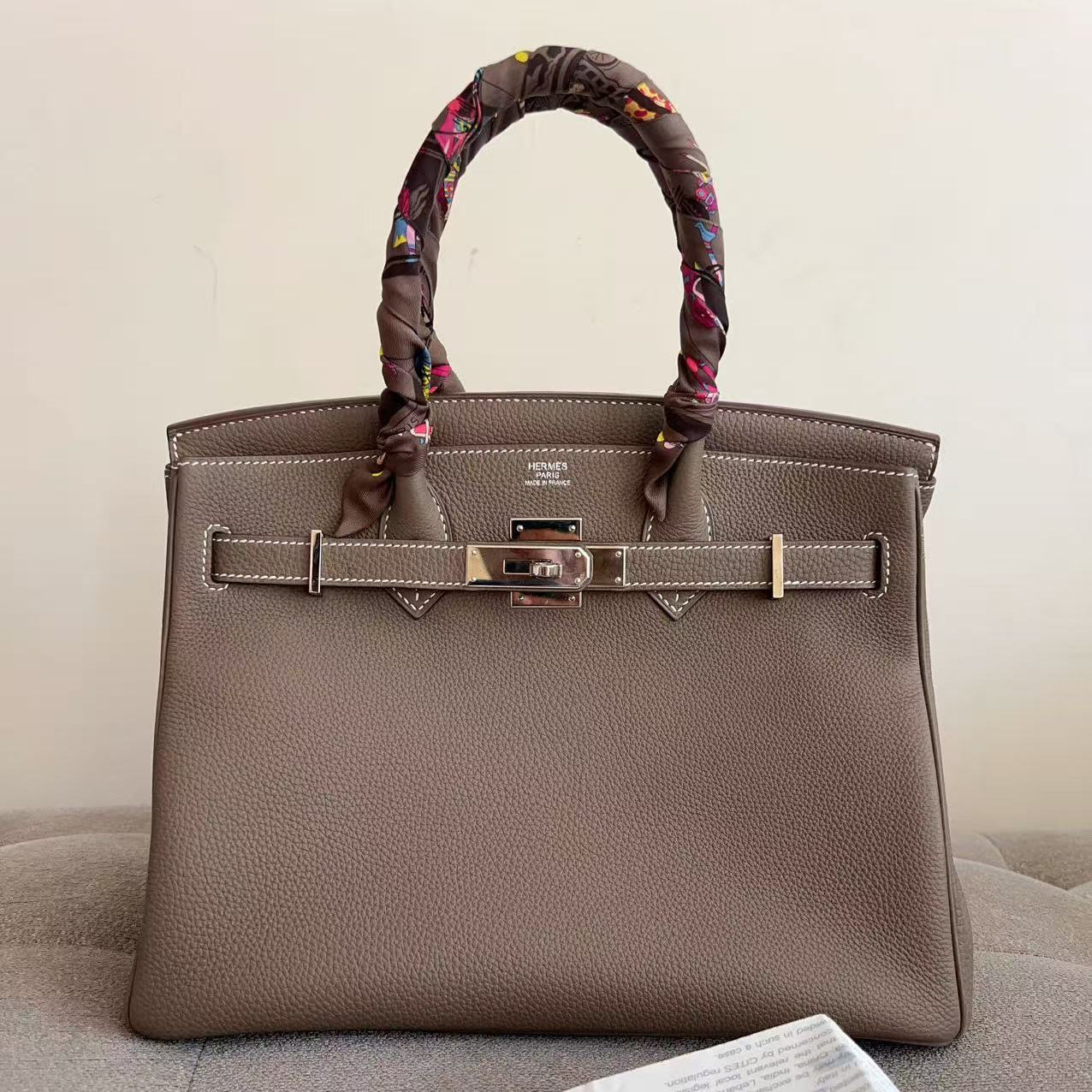 A taupe pebbled leather handbag with a matching leather strap handle wrapped in a colorful patterned scarf, resting on a gray tufted sofa with a paper document in the foreground.