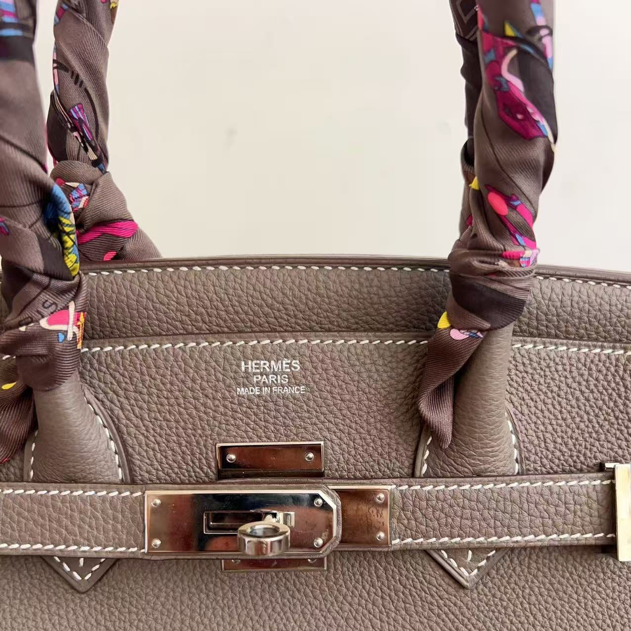 Close-up of a taupe pebbled leather handbag with silver-toned hardware and white contrast stitching, featuring a patterned brown silk scarf tied to the handles and visible HERMES PARIS branding on the front.