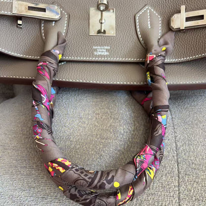 A close-up of a taupe leather handbag with silver-tone hardware and a brown silk scarf wrapped around the handle, set against a gray textured fabric background.