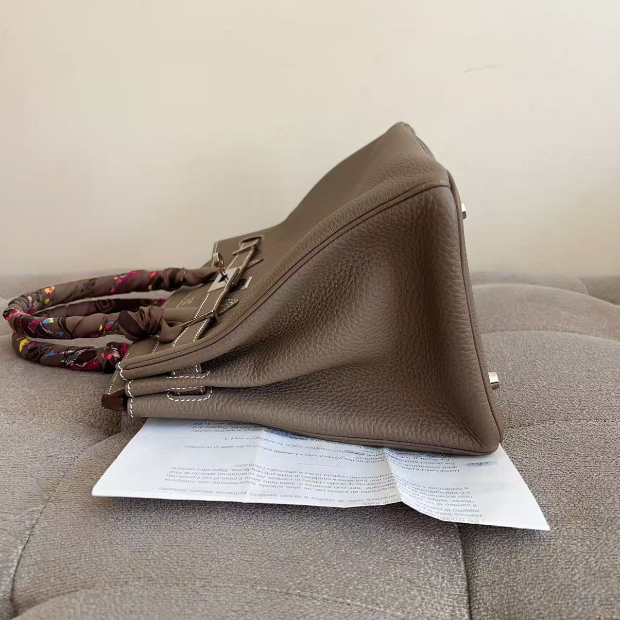 A taupe brown pebbled leather handbag with a patterned fabric strap, resting on a gray tufted sofa with a white paper sheet underneath.
