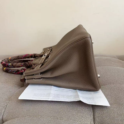 A taupe brown pebbled leather handbag with a patterned fabric strap, resting on a gray tufted sofa with a white paper sheet underneath.