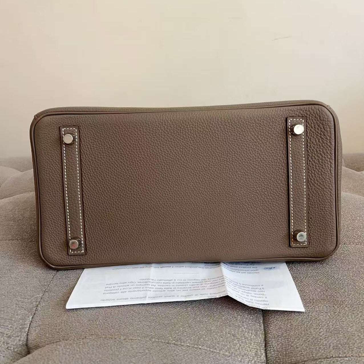 A taupe brown pebbled leather clutch or wallet shown from the bottom, resting on a tufted beige sofa with a paper insert underneath. The bag features silver-tone metal studs and contrast stitching.
