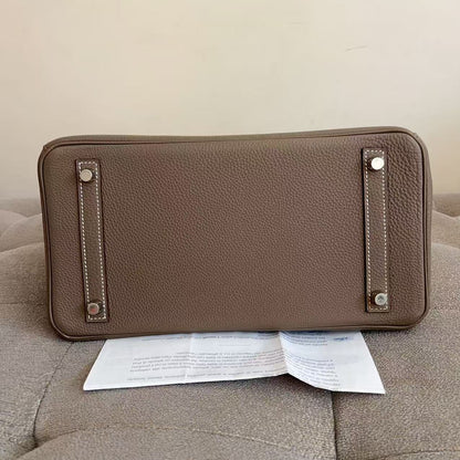 A taupe brown pebbled leather clutch or wallet shown from the bottom, resting on a tufted beige sofa with a paper insert underneath. The bag features silver-tone metal studs and contrast stitching.