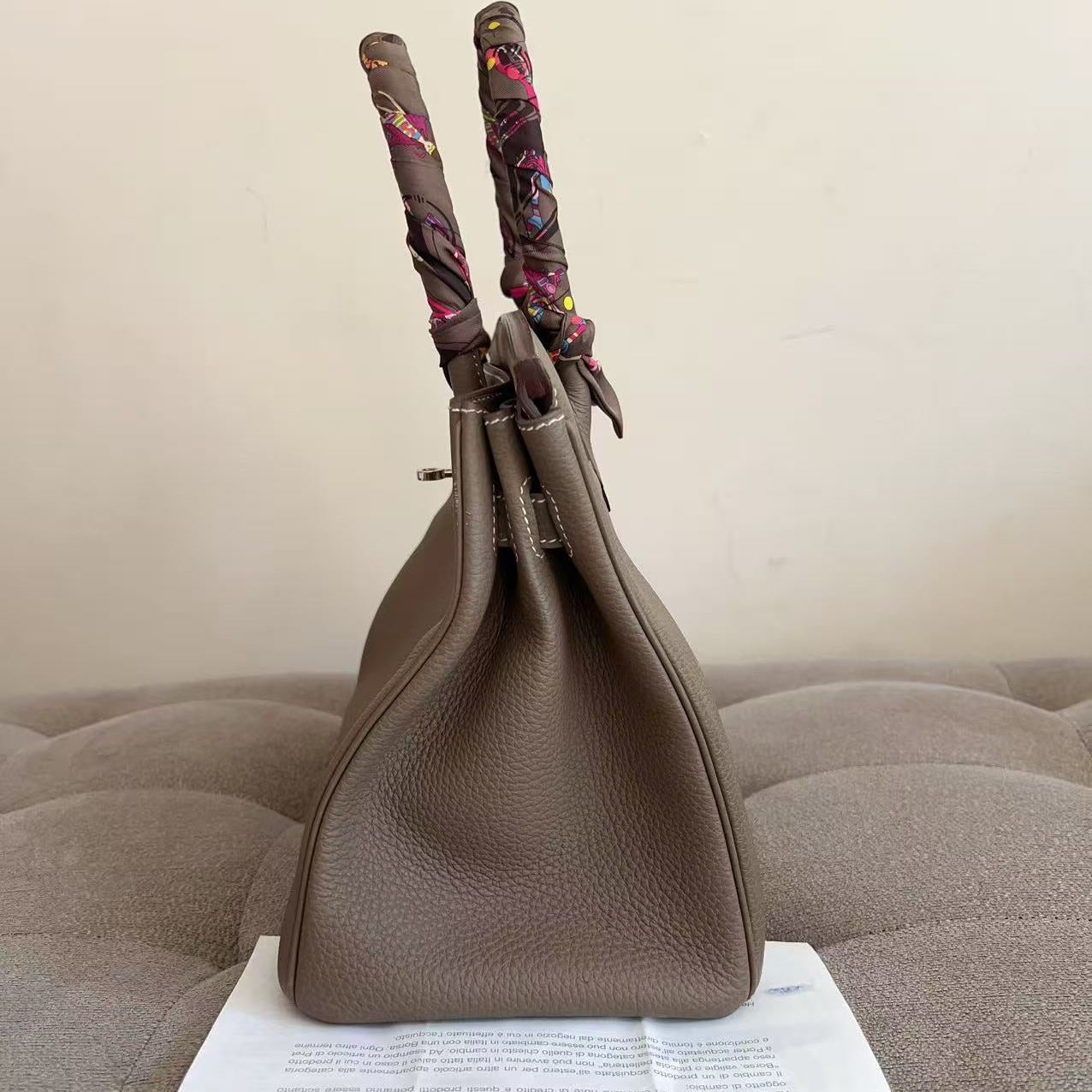 A taupe brown pebbled leather handbag with patterned scarf handles, standing on a white paper sheet placed on a tufted gray sofa against a plain beige wall.