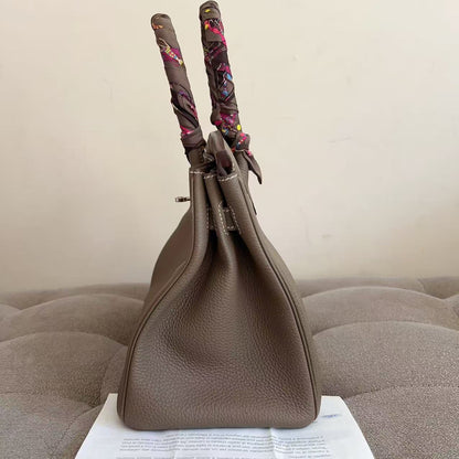 A taupe brown pebbled leather handbag with patterned scarf handles, standing on a white paper sheet placed on a tufted gray sofa against a plain beige wall.