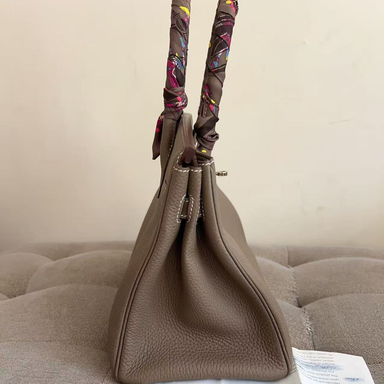 A taupe brown pebbled leather handbag with patterned scarf-wrapped handles, displayed on a tufted gray sofa with an instruction sheet in front.