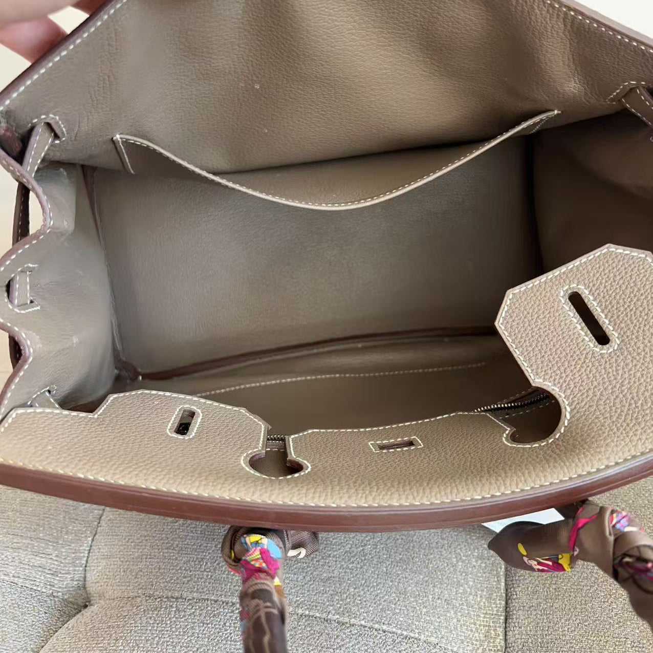 Close-up of the interior of a taupe leather handbag held open by a hand, showing the lining, interior pocket, and hardware details.