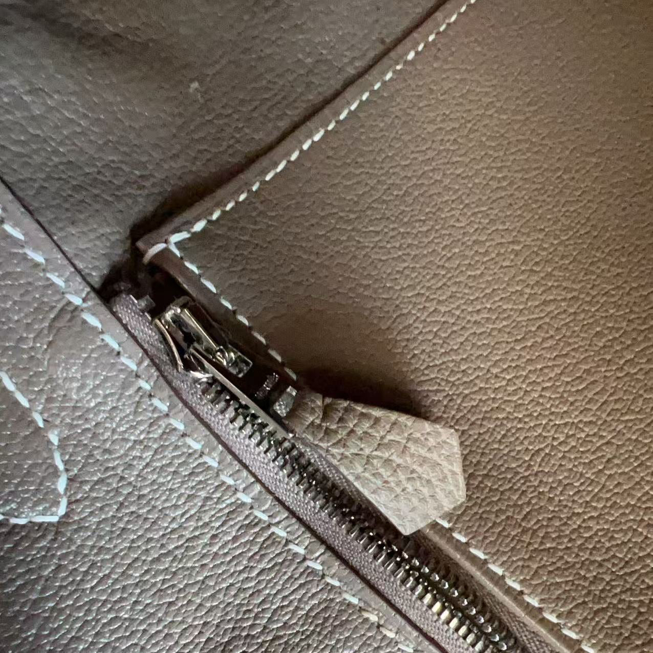 Close-up of a pebbled leather zipper detail on a taupe-brown bag, showing a silver-toned metal zipper pull with a matching leather tab and white contrast stitching.