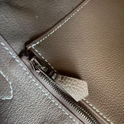 Close-up of a pebbled leather zipper detail on a taupe-brown bag, showing a silver-toned metal zipper pull with a matching leather tab and white contrast stitching.