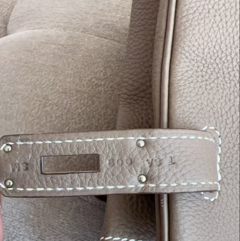 A close-up of a hand holding a taupe leather strap tag with white stitching and silver-tone rivets, attached to a matching taupe leather handbag.