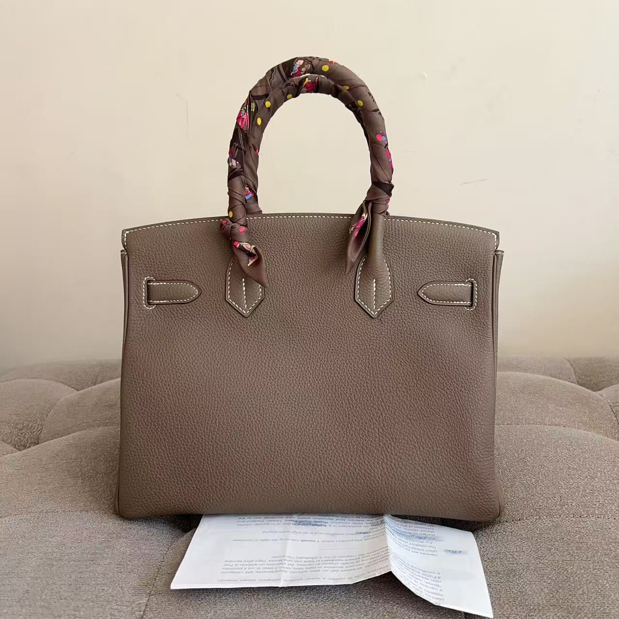 A taupe brown pebbled leather handbag with a matching fabric scarf wrapped around its top handles, displayed on a tufted beige sofa with a paper insert underneath.