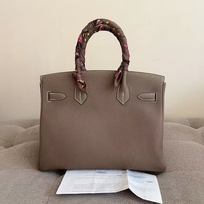 A taupe brown pebbled leather handbag with a matching fabric scarf wrapped around its top handles, displayed on a tufted beige sofa with a paper insert underneath.