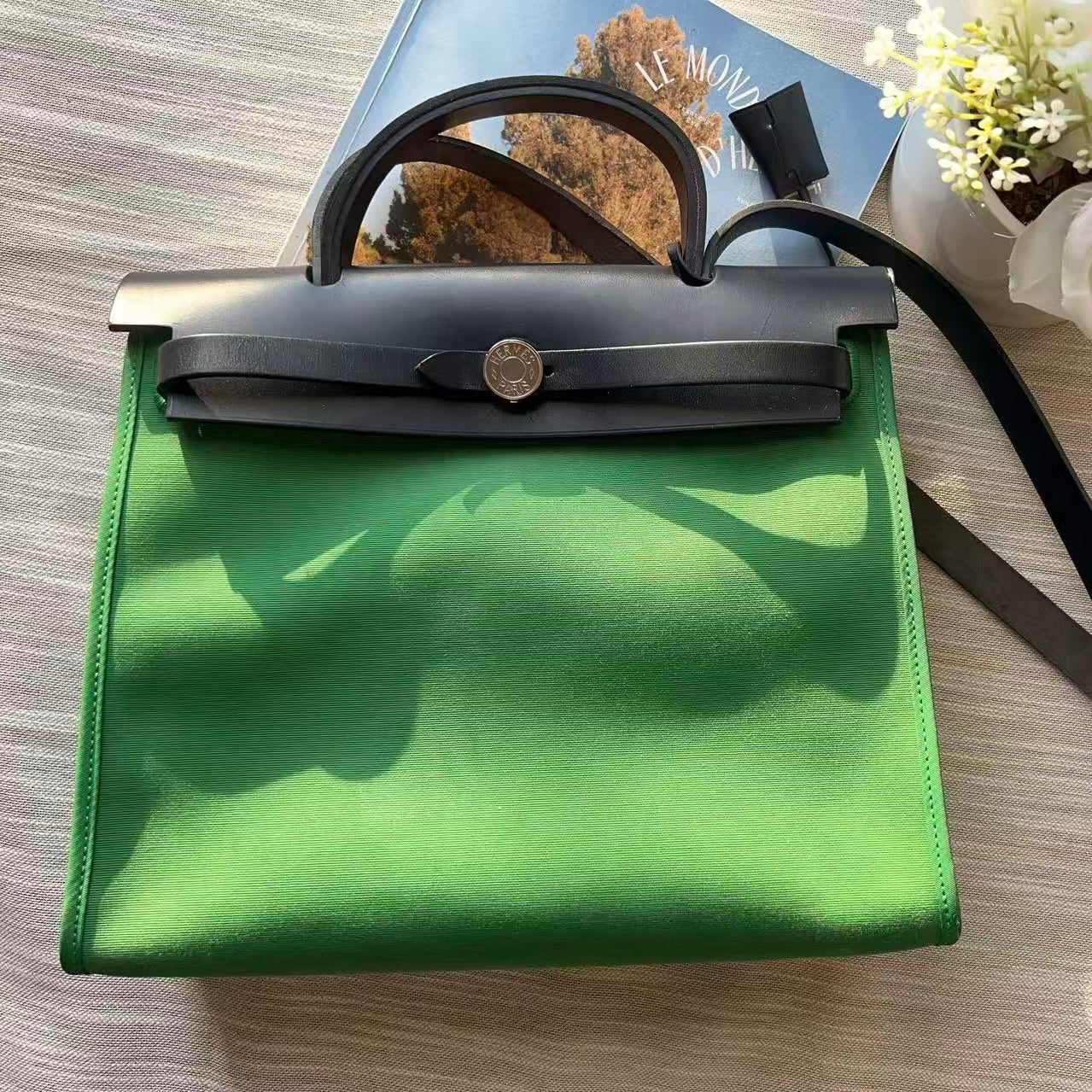 Green handbag with black handle on a wooden surface with flowers and magazine in the background