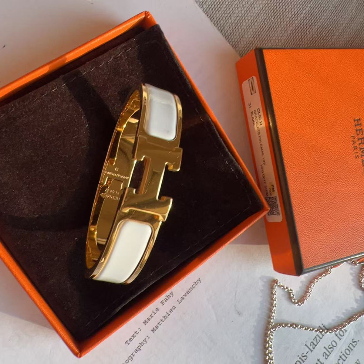 A gold-tone H-shaped cuff bracelet with white inlay, presented in an orange Hermes box next to the orange Hermes packaging and a silver chain.