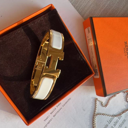 A gold-tone H-shaped cuff bracelet with white inlay, presented in an orange Hermes box next to the orange Hermes packaging and a silver chain.