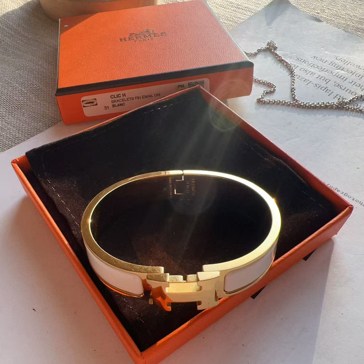 A gold-tone H-ear cuff bracelet with white inlay, presented in an orange Hermes box on a white surface with the orange Hermes box and chain in the background.