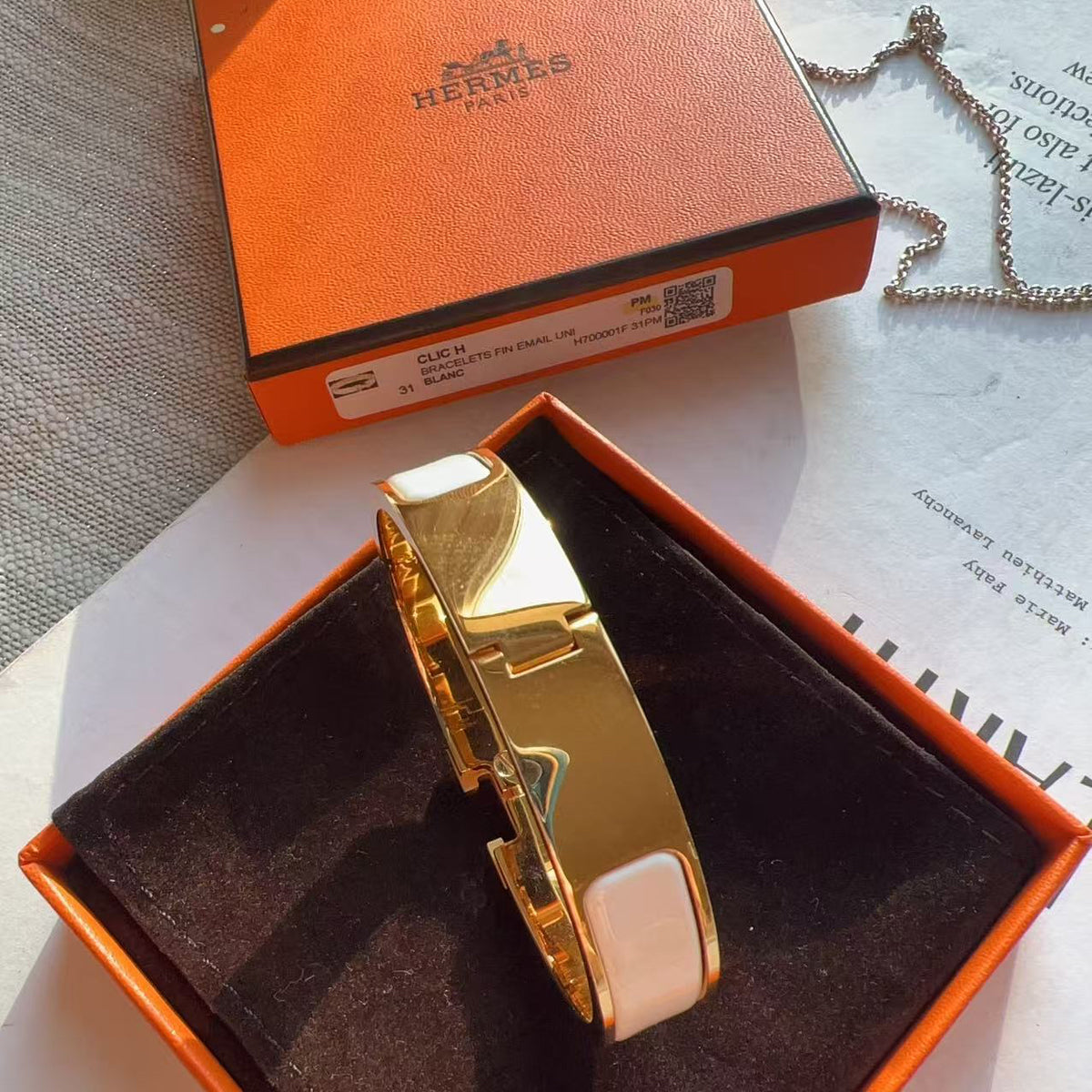 A gold-toned Hermès bracelet with white inlays, presented in an orange Hermès box on a white surface with the orange box lid in the background.