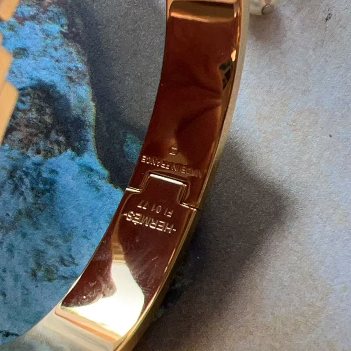 Close-up of a polished gold-toned metal bracelet or bangle clasp area, showing engraved text including 'HERMES' and '5107', resting on a textured surface with a blue-green stone prop.