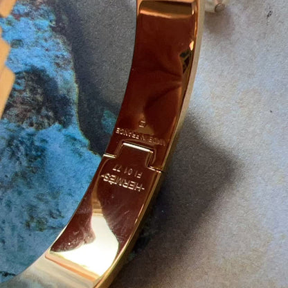 Close-up of a polished gold-toned metal bracelet or bangle clasp area, showing engraved text including 'HERMES' and '5107', resting on a textured surface with a blue-green stone prop.