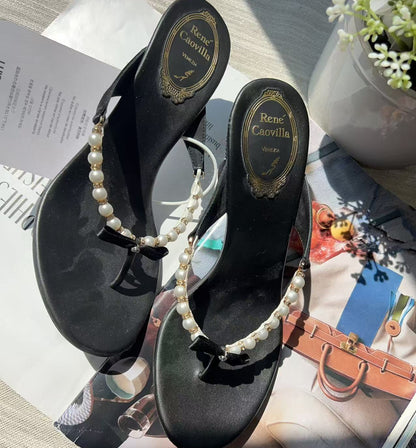 A pair of black thong sandals with pearl straps and small black bow details, displayed on top of a magazine page with a small potted plant in the background.