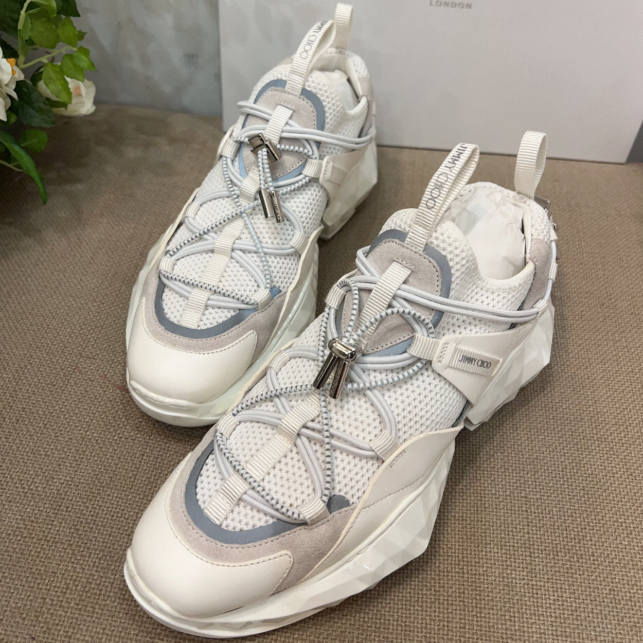 White sneakers with blue accents on a textured surface, with a Jimmy Choo box in the background.
