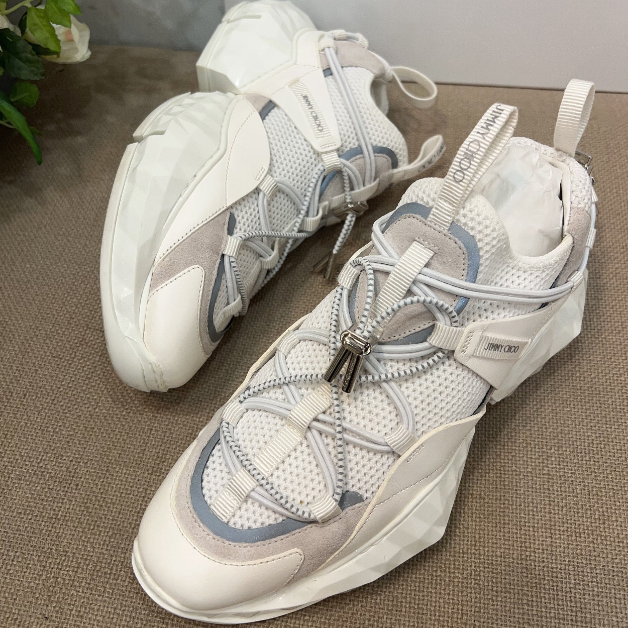 White and gray sneakers on a textured surface with 'Jimmy Choo' branding in the background.