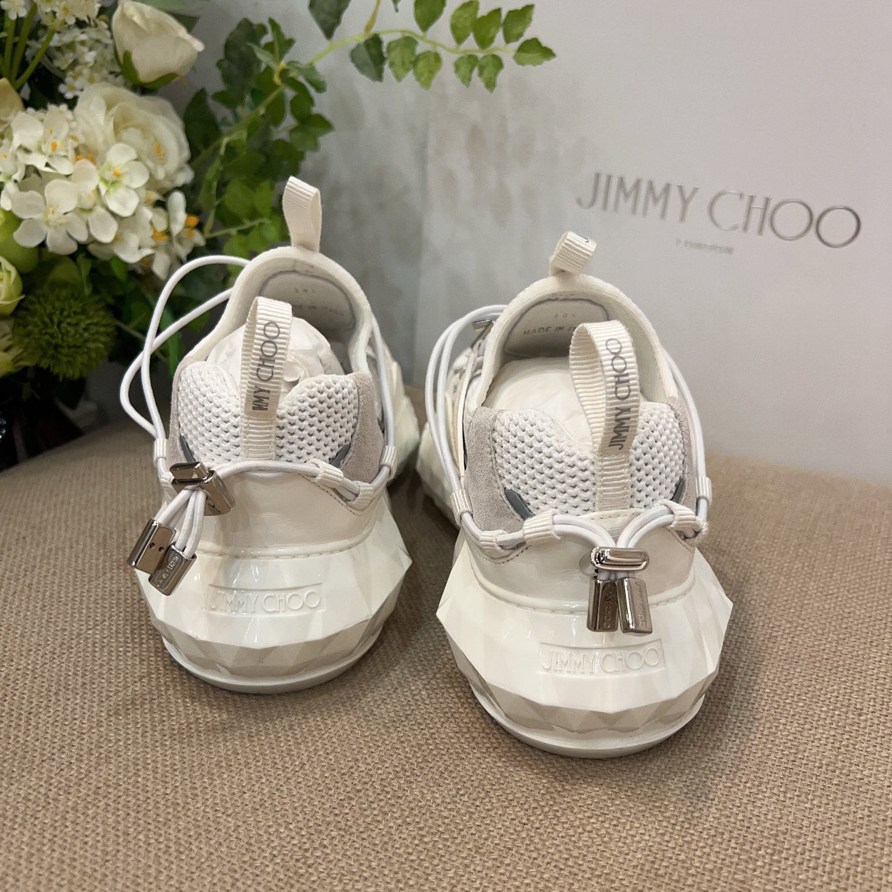 White sneakers with silver accents on a textured surface, with 'Jimmy Choo' branding in the background.