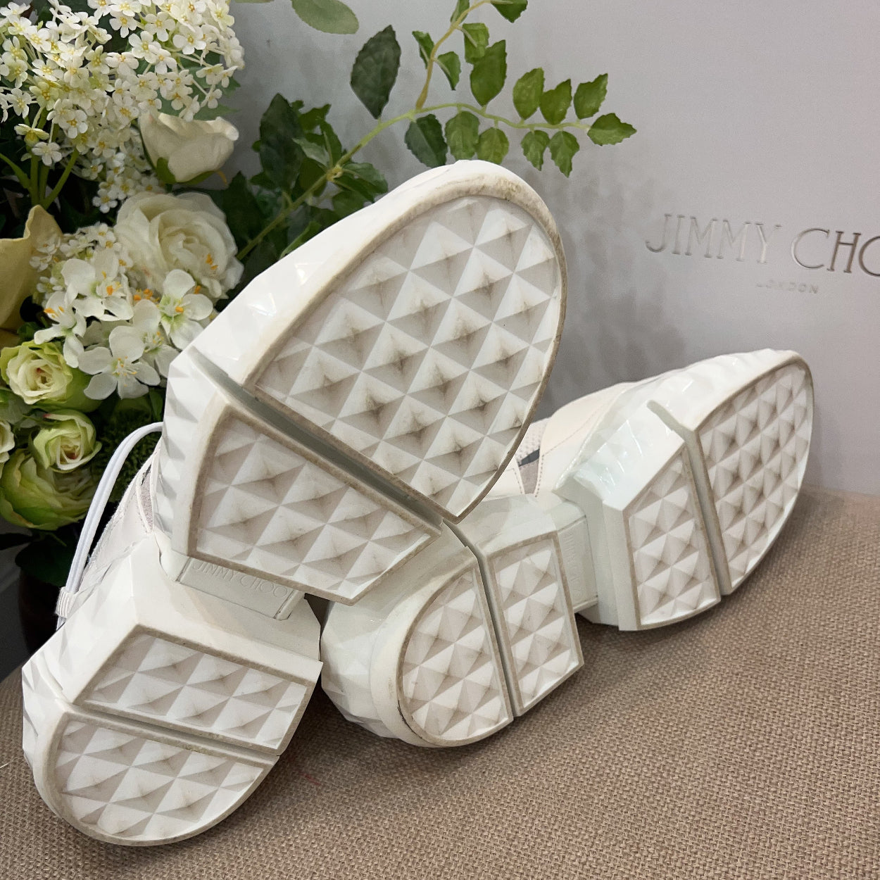 White sandals with textured sole on a brown surface, with 'Jimmy Choo' branding in the background.